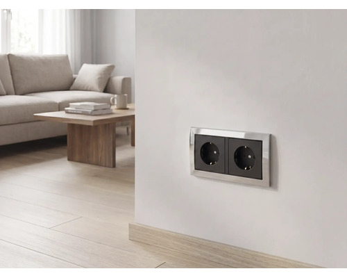 Zweifache Schutzkontakt-Steckdose mit schwarzen Einsätzen und silbernem Rahmen an einer Wand in einem hellen Wohnzimmer.