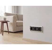 Dreifach-Steckdose mit Metallrahmen in einem hellen Wohnzimmer mit Sofa, Holztisch und Parkettboden.