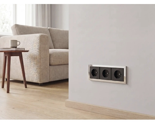 Dreifach-Steckdose mit Metallrahmen in einem hellen Wohnzimmer mit Sofa, Holztisch und Parkettboden.