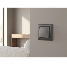 Anthrazitfarbener Lichtschalter an einer grauen Wand in einem modern eingerichteten Wohnzimmer mit Sofa und Holztisch.