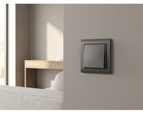 Anthrazitfarbener Lichtschalter an einer grauen Wand in einem modern eingerichteten Wohnzimmer mit Sofa und Holztisch.