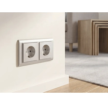 Doppelsteckdose an einer hellen Wand in einem modern eingerichteten Wohnzimmer mit Holztisch und Teppich.