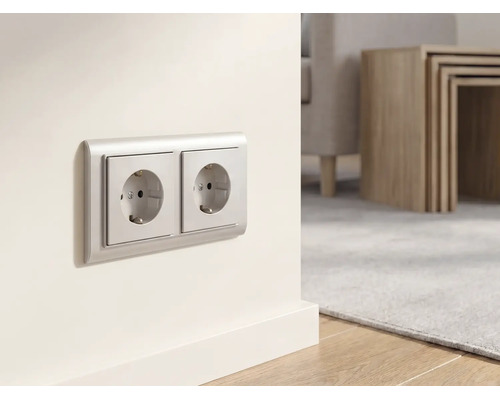 Doppelsteckdose an einer hellen Wand in einem modern eingerichteten Wohnzimmer mit Holztisch und Teppich.