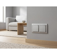 Moderner silberner Zweifach-Lichtschalter an einer grauen Wand in einem hellen Wohnzimmer mit Sofa und Holztisch.