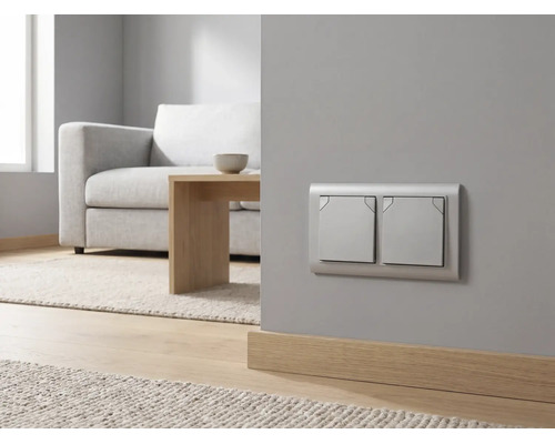 Moderner silberner Zweifach-Lichtschalter an einer grauen Wand in einem hellen Wohnzimmer mit Sofa und Holztisch.