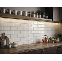 Küchenrückwand mit weißen, glänzenden Metro-Fliesen im Ziegelverband, beleuchtet durch LED-Licht unter einem Holzregal auf einer Holzplatte.