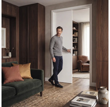 Ein Mann öffnet eine weiße Schiebetür in einem modernen Wohnzimmer mit dunklen Holzschränken, grünem Sofa und Holzfußboden.