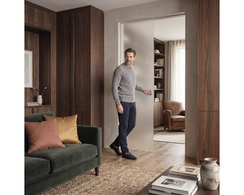 Ein Mann öffnet eine Schiebetür aus satiniertem Glas in einem modernen Wohnzimmer mit dunklen Holzschränken und einem grünen Samtsofa.