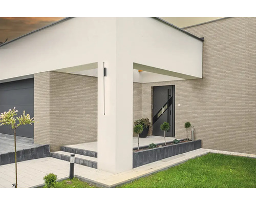 Moderne schwarze Außenwandleuchte an einem weißen Pfeiler, Hauseingang mit Klinkerfassade, Garage und Gartenbereich.