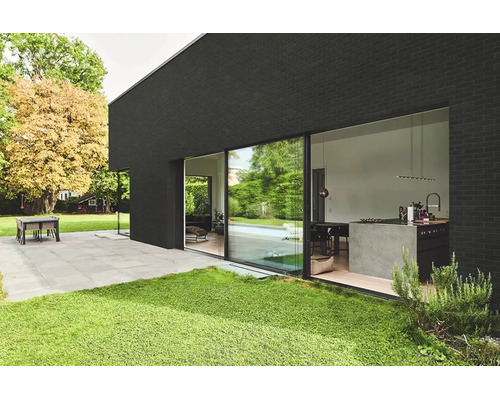 Moderne Hausfassade mit großer Glasschiebetür, Blick in die Küche, davor eine Terrasse mit Steinplatten und Rasenfläche.