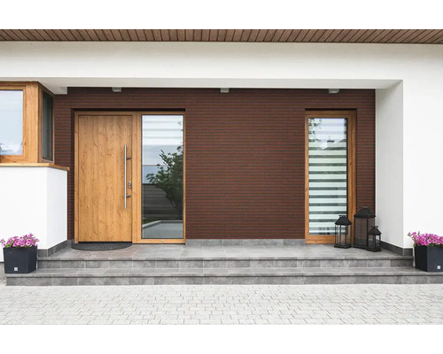 Moderner Hauseingang mit dunkelbrauner Klinkerfassade, Holztür, großen Fenstern, grauen Fliesenstufen und Blumenkübeln.