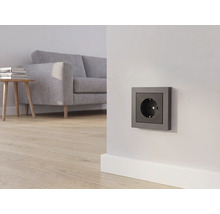 Anthrazitfarbene Steckdose an einer weißen Wand in einem hellen Wohnzimmer mit grauem Sofa und Holzboden.