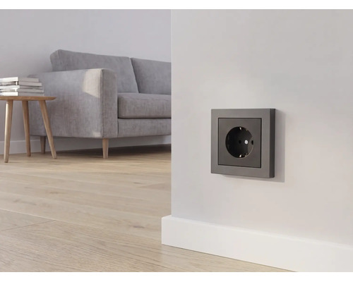 Anthrazitfarbene Steckdose an einer weißen Wand in einem hellen Wohnzimmer mit grauem Sofa und Holzboden.