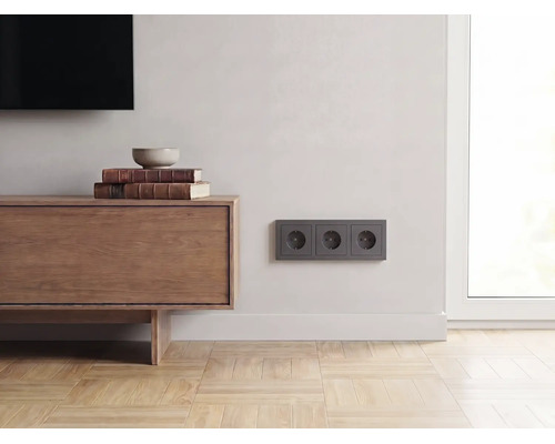 Dreifache Steckdose in Anthrazit an einer hellen Wand in einem modern eingerichteten Wohnzimmer mit Sideboard aus Holz und Parkettboden.