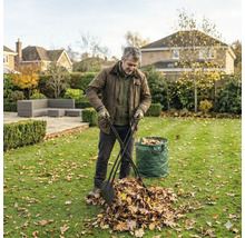 Ein Mann sammelt Herbstlaub mit einem Laubgreifer auf einem Rasen neben einem vollen Gartensack in einem weitläufigen Garten.