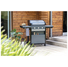 Grauer Campingaz Gasgrill mit drei Brennern auf einer hellen Holzterrasse vor einer Holzwand mit einer Pflanze.