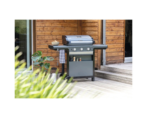 Grauer Campingaz Gasgrill mit drei Brennern auf einer hellen Holzterrasse vor einer Holzwand mit einer Pflanze.