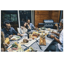 Personengruppe beim Grillabend auf einer Holzterrasse an einem gedeckten Tisch neben einem modernen Gasgrill.