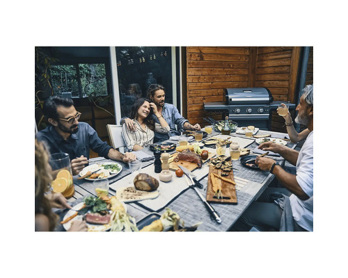 Personengruppe beim Grillabend auf einer Holzterrasse an einem gedeckten Tisch neben einem modernen Gasgrill.