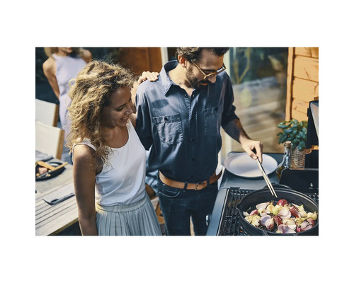 Ein Paar bereitet Gemüse in einer gusseisernen Pfanne auf einem Gasgrill im hellen Außenbereich einer Terrasse zu.