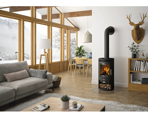 Moderner schwarzer Kaminofen mit brennendem Feuer in einem hellen Wohnzimmer mit Sofa, Holztisch und großen Panoramafenstern mit Schneeblick.