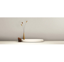 Minimalistische Szene mit einem weißen runden Podium, einer schlanken goldfarbenen Vase mit Dekozweigen und einem kleinen braunen Gittersessel vor einer strukturierten Wand.