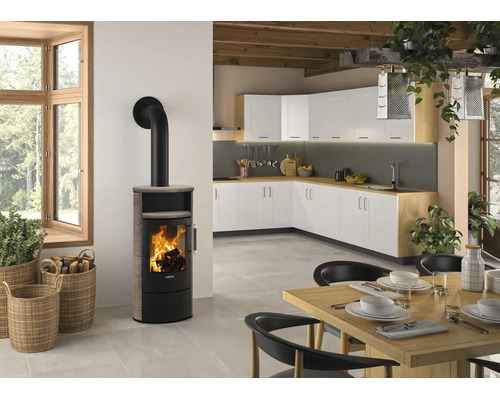 Moderner Kaminofen mit brennendem Feuer in einer hellen Wohnküche mit Küchenschränken, Holztisch und Weidenkörben für Brennholz.