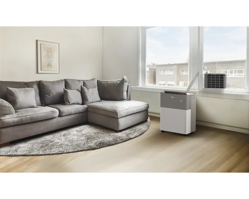 Mobiles Klimagerät in einem hellen Wohnzimmer mit grauem Ecksofa und Fensterdurchführung für den Abluftschlauch.