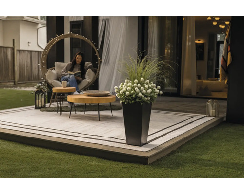 Moderne Terrasse mit Outdoorteppich, runden Holztischen, einem Hängesessel und einem hohen Pflanzgefäß mit Blumen und Gräsern.