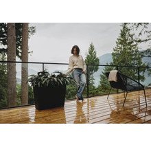 Moderne Holzterrasse mit einem großen schwarzen Pflanzgefäß, einem schwarzen Loungesessel und Glasgeländer vor einer Wald- und Bergkulisse.