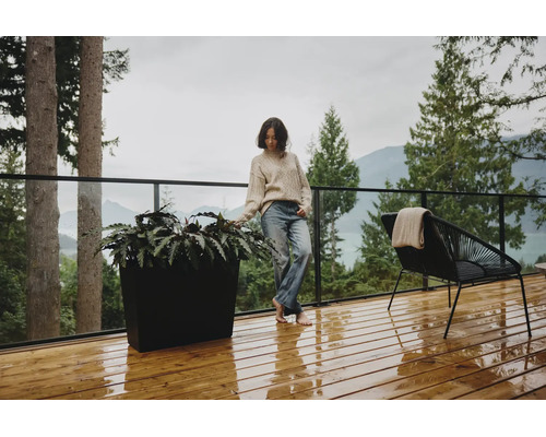 Moderne Holzterrasse mit einem großen schwarzen Pflanzgefäß, einem schwarzen Loungesessel und Glasgeländer vor einer Wald- und Bergkulisse.