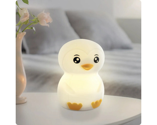 Pinguinförmiges Nachtlicht leuchtend auf einem Nachttisch neben einer Vase mit Rosen in einem hellen Schlafzimmer.