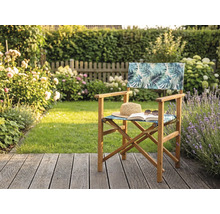 Regiestuhl aus Holz mit tropischem Blattmuster auf einer Garten-Holzterrasse mit Strohhut und Buch vor einer grünen Hecke.