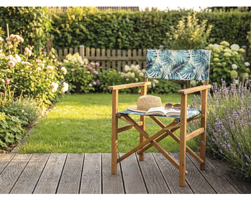 Regiestuhl aus Holz mit tropischem Blattmuster auf einer Garten-Holzterrasse mit Strohhut und Buch vor einer grünen Hecke.