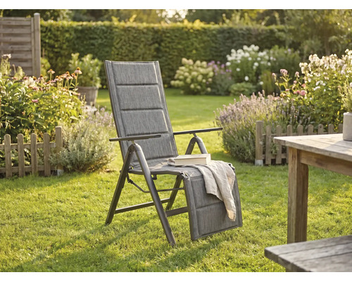 Gepolsterter Garten-Hochlehner mit verstellbarer Fußstütze in einem hellen Garten mit Rasen, Blumenbeeten und Holztisch.