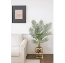 Künstliche Palme in einem Rattan-Pflanzenständer neben einem hellen Sofa in einem hellen Raum mit Holzboden.