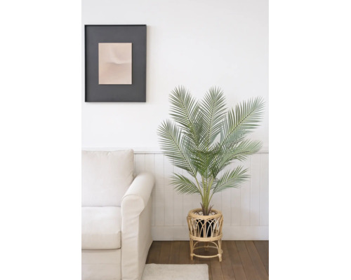 Künstliche Palme in einem Rattan-Pflanzenständer neben einem hellen Sofa in einem hellen Raum mit Holzboden.