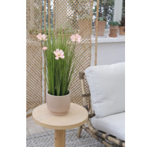 Künstliche Gräser mit rosa Blüten in einem geriffelten Übertopf auf einem runden Holztisch neben einem Sessel mit weißem Polster.