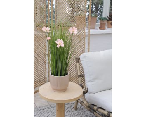 Künstliche Gräser mit rosa Blüten in einem geriffelten Übertopf auf einem runden Holztisch neben einem Sessel mit weißem Polster.