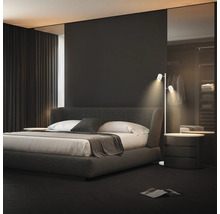 Modernes Schlafzimmer mit einem dunkelgrauen Polsterbett, weißer Bettwäsche und einer schmalen weißen Stehleuchte mit zwei Strahlern.