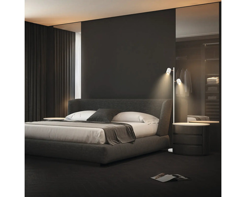 Modernes Schlafzimmer mit einem dunkelgrauen Polsterbett, weißer Bettwäsche und einer schmalen weißen Stehleuchte mit zwei Strahlern.