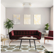 Modernes helles Wohnzimmer mit weinrotem Samtsofa, dreieckigem Couchtisch aus Holz, gemustertem Teppich, Wandbildern und Deckenleuchte.