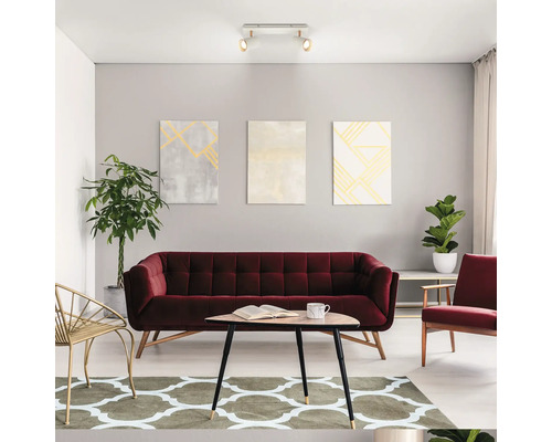 Modernes helles Wohnzimmer mit weinrotem Samtsofa, dreieckigem Couchtisch aus Holz, gemustertem Teppich, Wandbildern und Deckenleuchte.