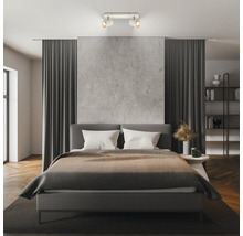 Modernes Schlafzimmer mit einem zweiflammigen Deckenstrahler, der ein graues Polsterbett und eine Wand in Betonoptik beleuchtet.