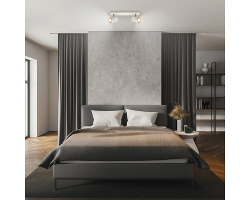 Modernes Schlafzimmer mit einem zweiflammigen Deckenstrahler, der ein graues Polsterbett und eine Wand in Betonoptik beleuchtet.