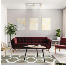 Modernes Wohnzimmer mit rotem Samtsofa, Holztisch, gemustertem Teppich, Zimmerpflanzen und einer Deckenleuchte mit drei verstellbaren Strahlern.