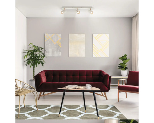 Modernes Wohnzimmer mit rotem Samtsofa, Holztisch, gemustertem Teppich, Zimmerpflanzen und einer Deckenleuchte mit drei verstellbaren Strahlern.