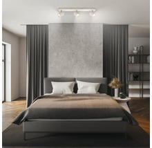 Modernes Schlafzimmer mit einem dreiflammigen Deckenstrahler, einem Polsterbett, einer Wand in Betonoptik und dunkelgrauen Vorhängen.