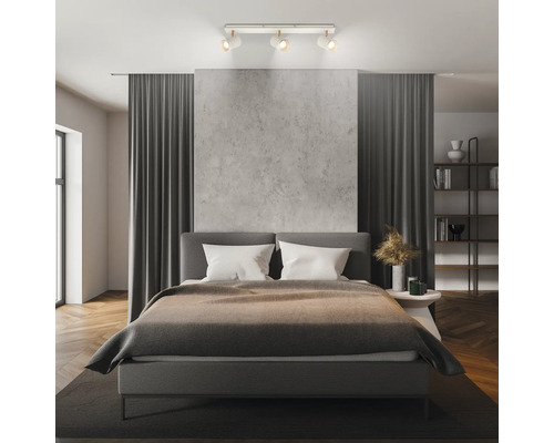 Modernes Schlafzimmer mit einem dreiflammigen Deckenstrahler, einem Polsterbett, einer Wand in Betonoptik und dunkelgrauen Vorhängen.