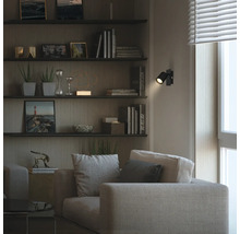 Modernes Wohnzimmer mit Stoffsofa und Wandregalen. Ein schwarzer verstellbarer LED-Wandstrahler erzeugt einen Lichtpunkt im dunklen Raum.